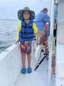 A young angler proudly displaying a caught fish while wearing a life jacket and colorful shorts on a fishing boat.