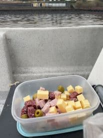 A container of assorted snacks including cheese, salami, and olives on a boat, with a view of the water in the background.