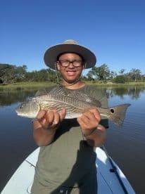 A young angler proudly displaying a striped bass while fishing on a calm river, surrounded by lush greenery.