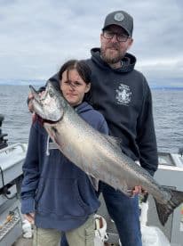 A young angler and an adult proudly displaying a large Salmon while fishing on a boat, showcasing a successful day on the water.