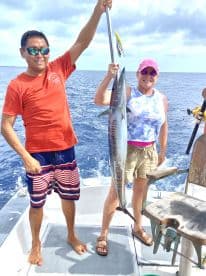 Two anglers proudly displaying a large fish, likely a Mackerel, while fishing offshore in a sunny environment.