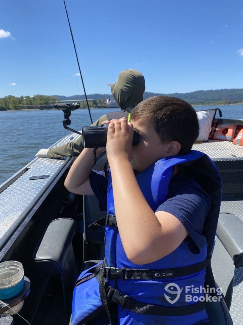 A young boy in a blue life jacket using binoculars while on a fishing boat, enjoying a sunny day on the water.