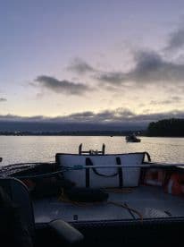 Interior view of a fishing boat at dawn, showcasing the seating and gear setup, with another boat visible in the distance on calm waters.