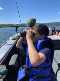 A young boy in a blue life jacket using binoculars while on a fishing boat, enjoying a sunny day on the water.