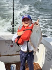 A young angler proudly displaying a Flounder while wearing a life jacket on a boat, showcasing a fun day of fishing.