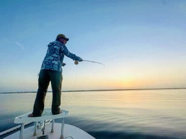 An angler casting a fly rod from the bow of a boat during a serene sunset on calm waters.
