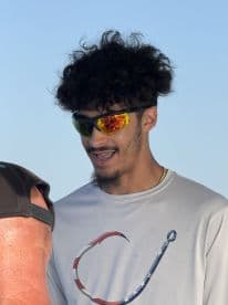 A young man wearing sunglasses and a fishing-themed shirt, engaged in conversation on a boat.