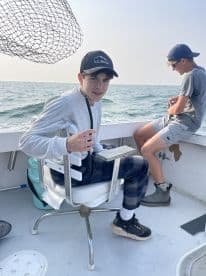 Two young anglers aboard a fishing boat, one seated in a chair with a fishing rod, while the other prepares to cast. The setting captures the excitement of a day on the water.