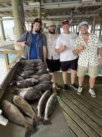 A group of four anglers proudly posing with their impressive catch of various fish, including speckled trout and black drum, displayed on a board at a fishing dock.