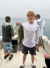 A young angler proudly displaying a large Largemouth Bass while fishing on a boat, with friends in the background enjoying the day on the water.