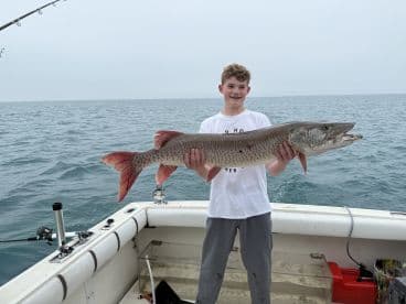 Young angler proudly holding a large Muskellunge while fishing on a boat in open waters.
