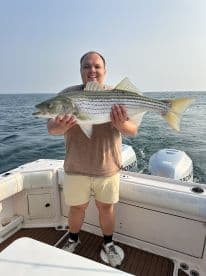 Angler proudly displaying a large Striped Bass while aboard a fishing boat, showcasing a successful day on the water.