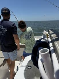 A young angler and an adult are engaged in fishing on a boat, with the child actively reeling in a catch in a sunny coastal environment.