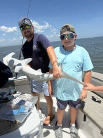 A young angler and an adult proudly displaying a caught shark on a fishing boat, showcasing a successful day on the water.