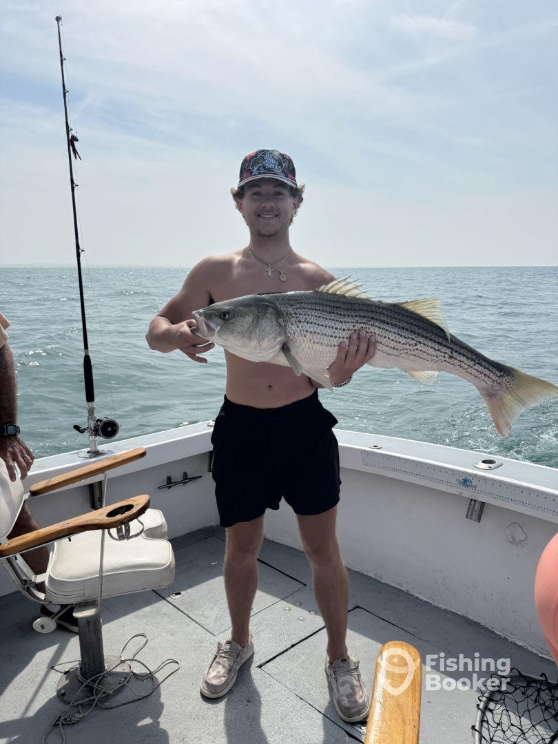 A young angler proudly displaying a large Striped Bass while fishing on a boat, showcasing a successful day on the water.