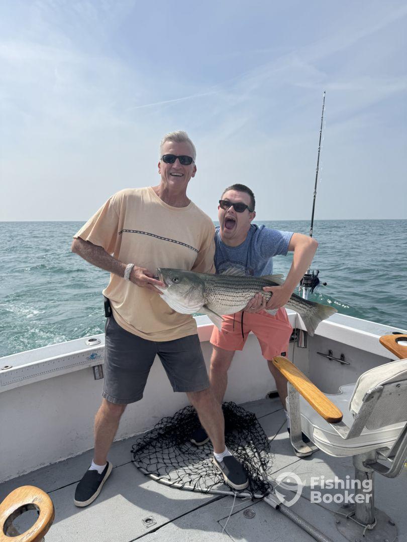 Two anglers celebrating a successful catch of a large striped bass while fishing on a charter boat.
