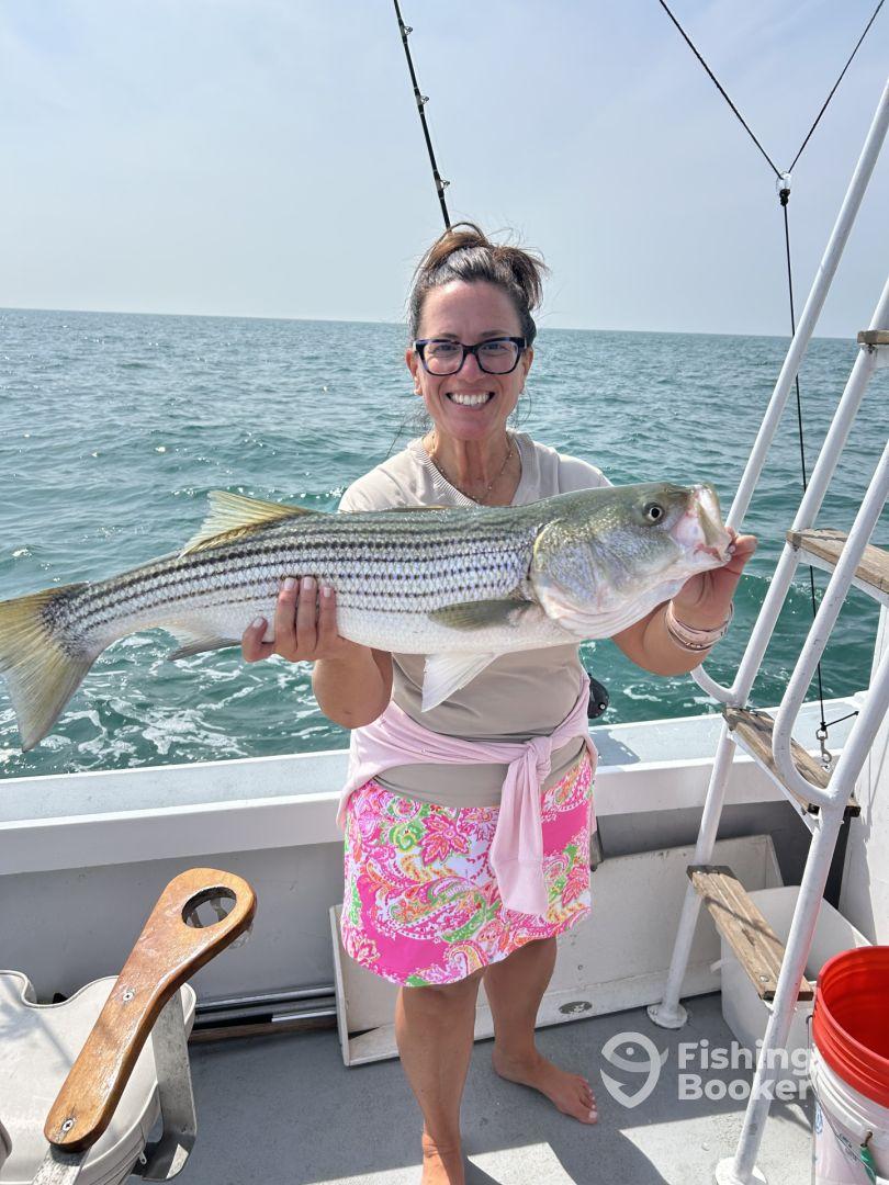An angler proudly displaying a large Striped Bass while fishing on a boat, showcasing a successful day on the water.