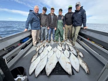A group of anglers proudly displaying their catch of various fish, including halibut and salmon, on the deck of a fishing boat in open waters.