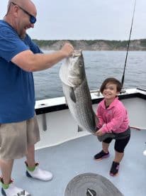 A young girl joyfully holds a large Striped Bass with the help of an adult on a fishing boat, showcasing a memorable catch during their fishing adventure.