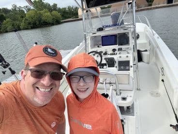 A father and son duo smiling for a selfie inside a fishing boat, showcasing the helm and seating area, ready for a day on the water.