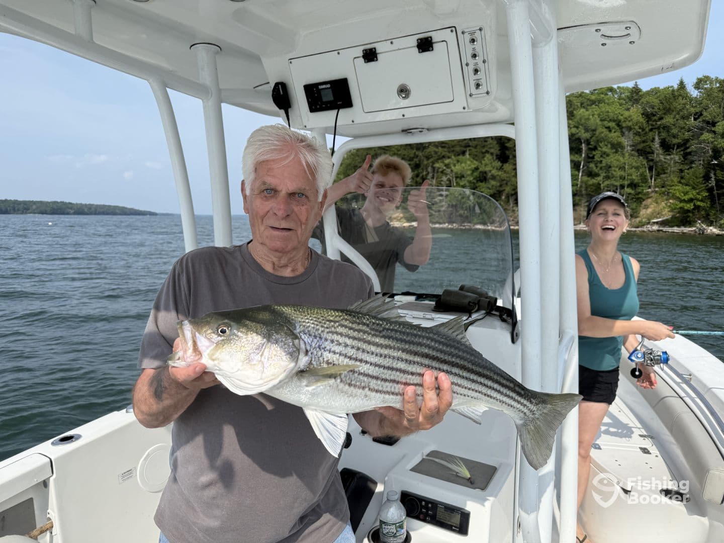 An angler proudly displaying a large striped bass aboard a boat, with fellow anglers enjoying the fishing experience in a scenic lake setting.