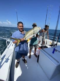 A young angler proudly displaying a large Yellowtail on a fishing boat, surrounded by fellow anglers enjoying a day on the water.