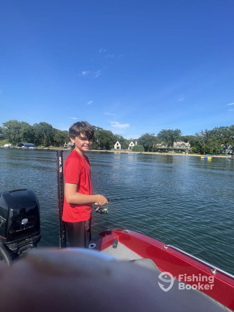 A young angler casting a line from a red boat on a calm lake, surrounded by scenic homes and greenery.