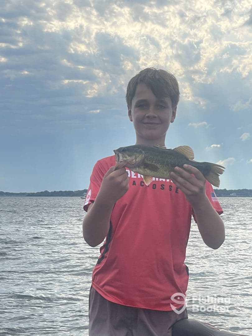 A young angler proudly displaying a large Largemouth Bass while fishing on a lake, showcasing a beautiful cloudy sky in the background.