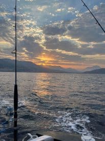 A picturesque sunset view from the side of a fishing boat, showcasing the tranquil waters and mountainous backdrop, perfect for a relaxing fishing experience.