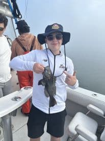A young angler proudly displaying a Black Sea Bass while fishing on a boat, surrounded by friends on a foggy day.