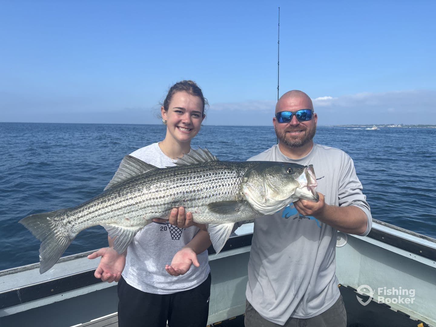 A young angler and an adult proudly displaying a large Striped Bass while fishing on a boat in open waters.