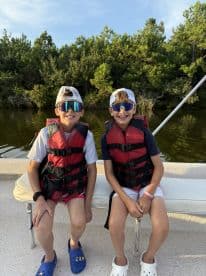 Two young boys wearing life jackets and sunglasses, enjoying a day on a boat in a scenic waterway.