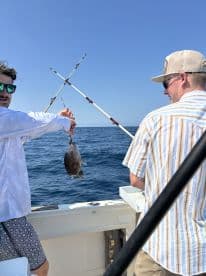 Two anglers on a boat enjoying a fishing trip, with one proudly displaying a freshly caught fish against a clear blue ocean backdrop.