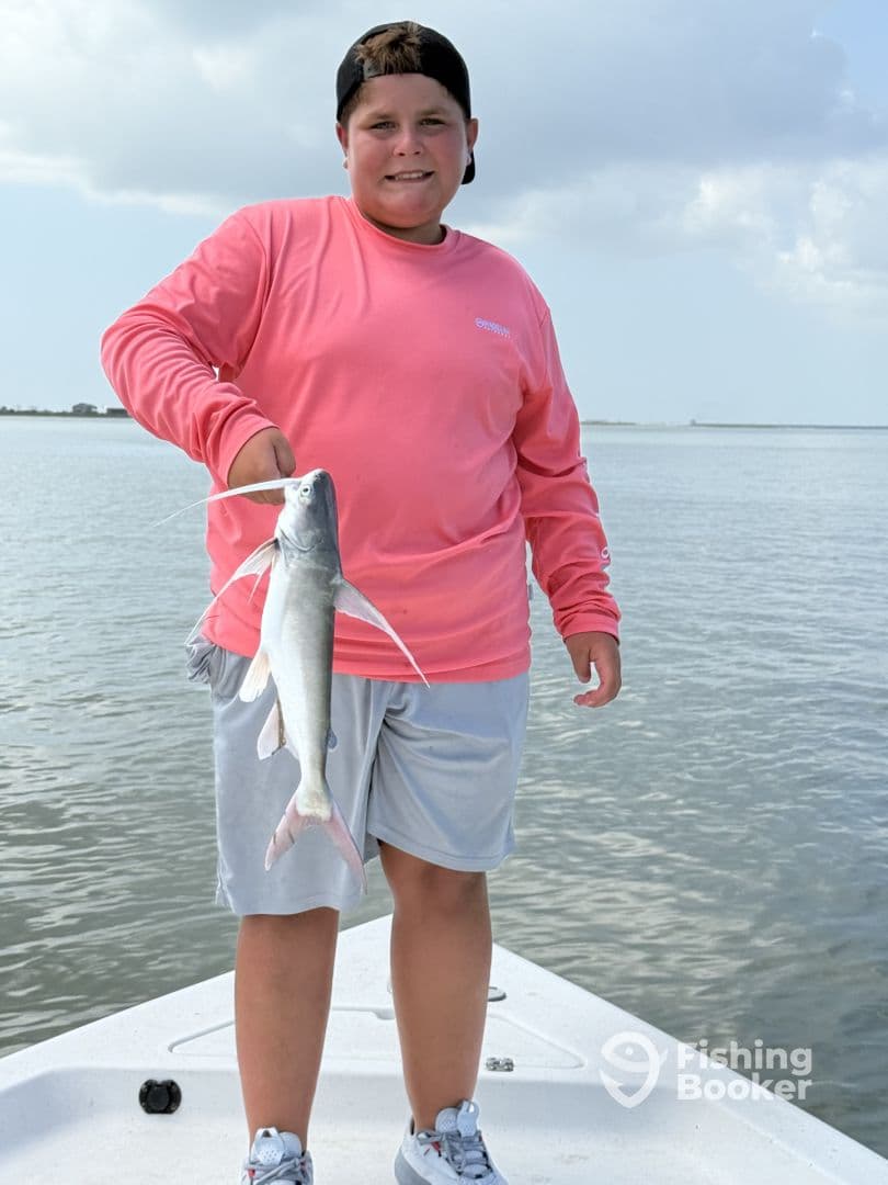 A young angler proudly displaying a catfish while standing on a boat in a calm fishing environment.
