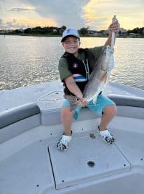 A young angler proudly displaying a large Redfish while fishing on a boat during sunset.