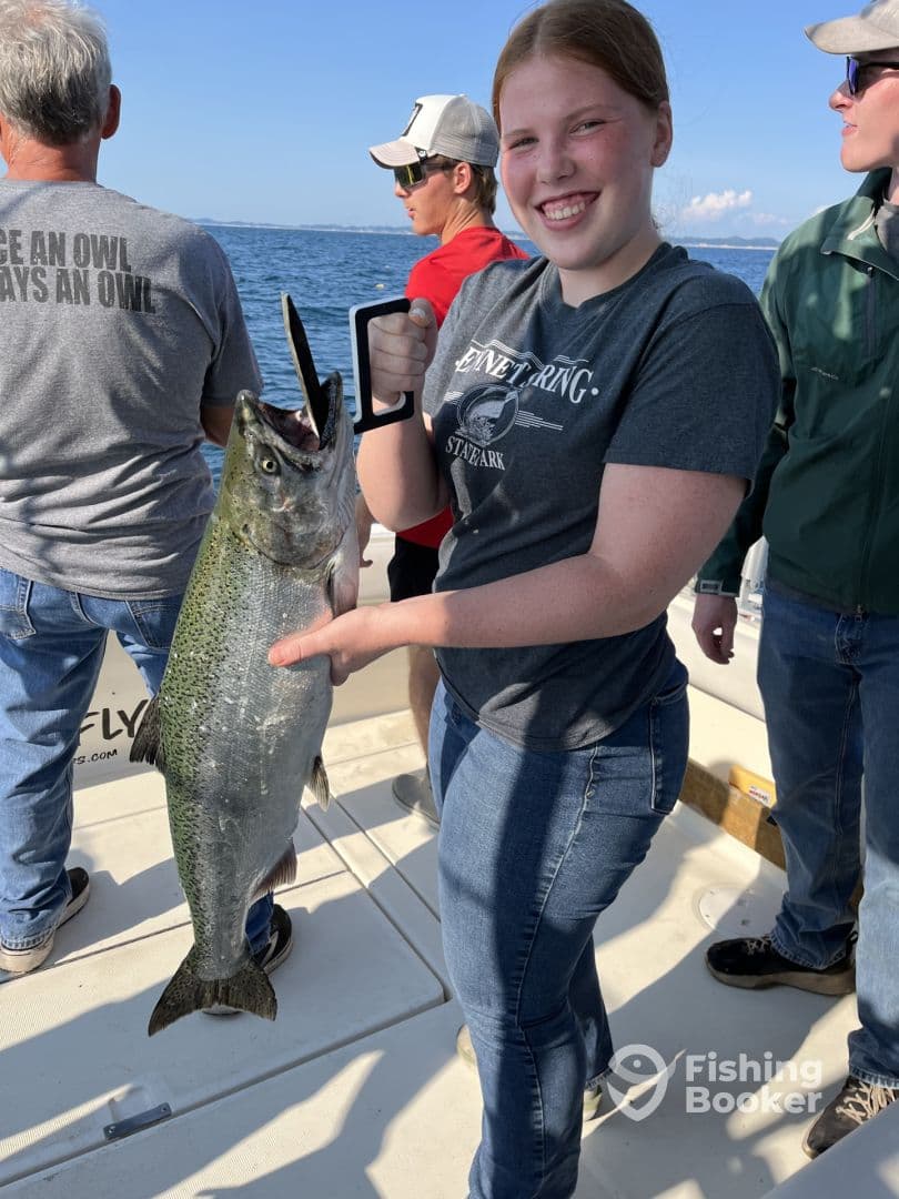 A young angler proudly displaying a large Salmon while fishing on a boat, surrounded by fellow anglers on a sunny day.