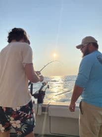 Two anglers engaged in fishing on a boat during sunset, with one reeling in a catch while the other observes.
