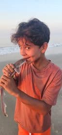 A young angler joyfully holding a small shark at the beach during sunset, showcasing a memorable fishing experience.