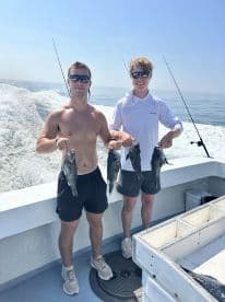 Two young anglers proudly displaying their catch of Black Sea Bass while on a fishing charter in open waters.