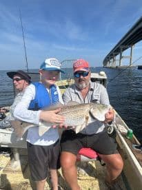 A young angler proudly displaying a large Redfish alongside an adult on a fishing boat, with a scenic bridge in the background.