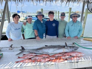 A group of anglers proudly displaying their catch, including a large King Mackerel and several Red Snapper, at a dockside cleaning station.