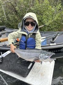 A young angler proudly displaying a large salmon while fishing from a boat in a lush, green environment.