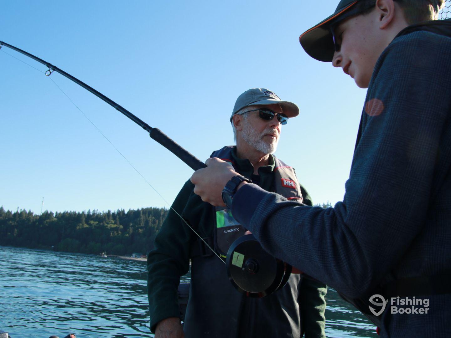 A young angler is reeling in a fish while being guided by an experienced fisherman on a tranquil lake, surrounded by lush greenery.