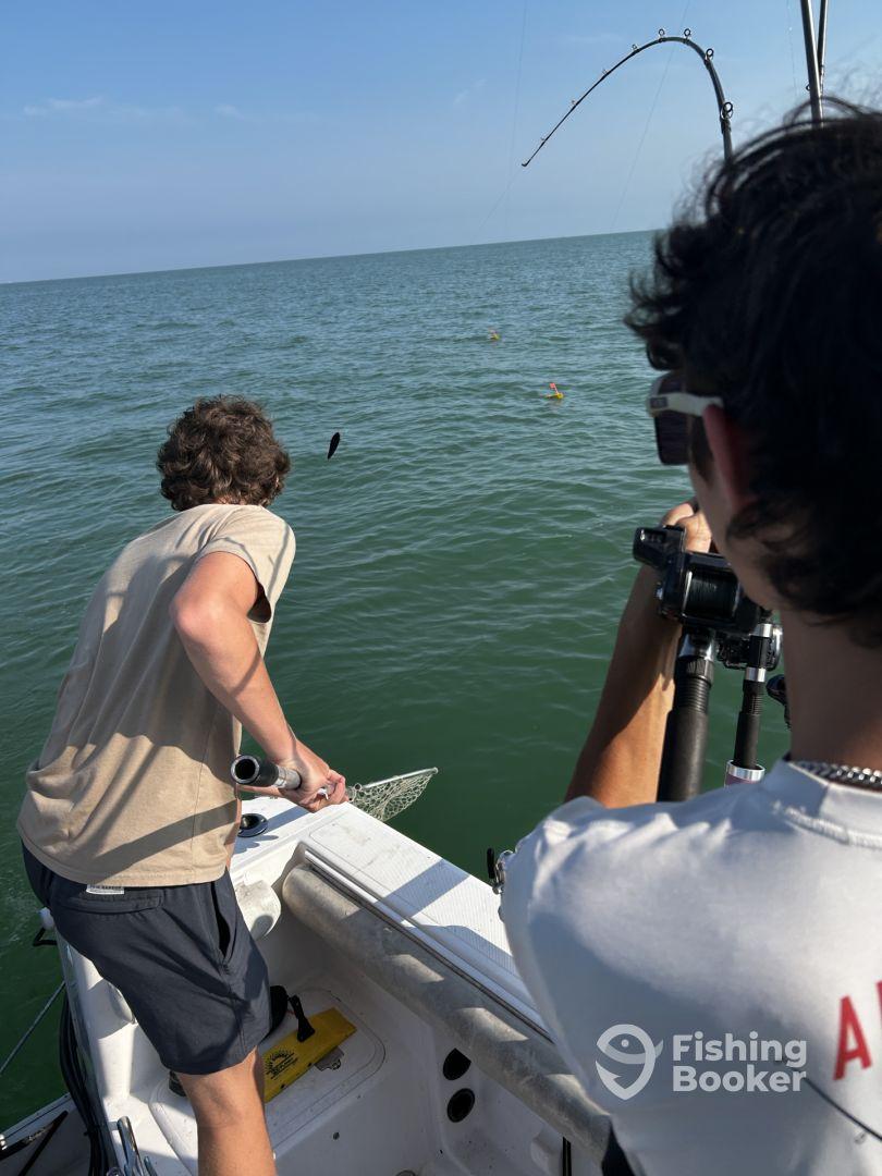 Two young anglers actively fishing from a boat, with one preparing to net a catch while the other captures the moment on camera.