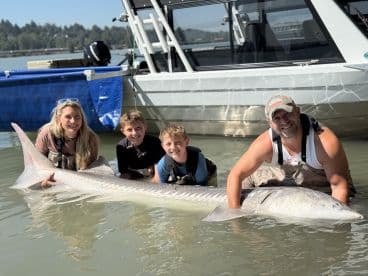A family proudly displays a large Sturgeon while wading in the water during a fishing trip, showcasing a memorable catch.