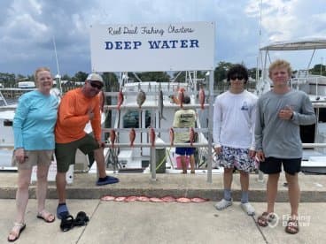 A group of four anglers proudly posing in front of their catch display, featuring several Red Snapper, at a marina after a successful fishing trip.