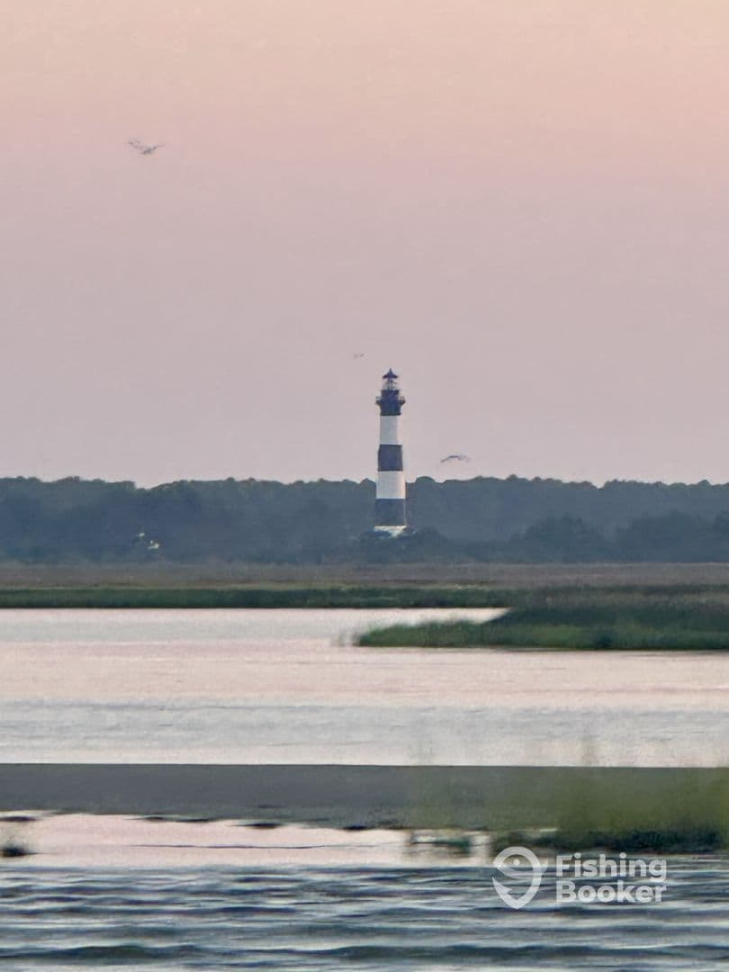 A distant view of a lighthouse against a pastel sky, with water and marshland in the foreground.