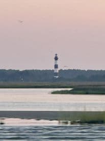 A distant view of a lighthouse against a pastel sky, with water and marshland in the foreground.