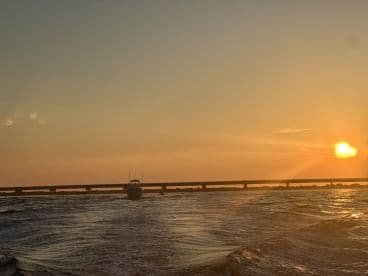 A scenic sunset view of a fishing boat navigating through calm waters, with a bridge in the background, capturing the tranquil atmosphere of a fishing trip.
