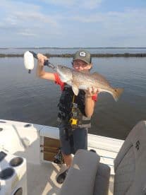 A young angler proudly displaying a large Redfish while fishing from a boat in calm waters.